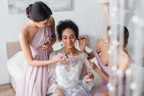young women doing makeup to happy african american bride in bedroom