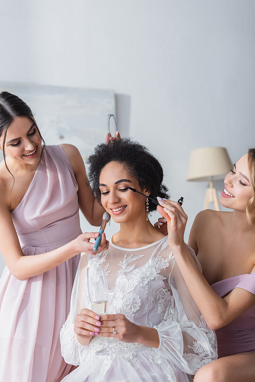 smiling bridesmaids applying mascara and face powder on african american bride