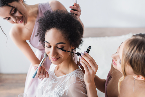 smiling women doing makeup to pretty african american bride, blurred background