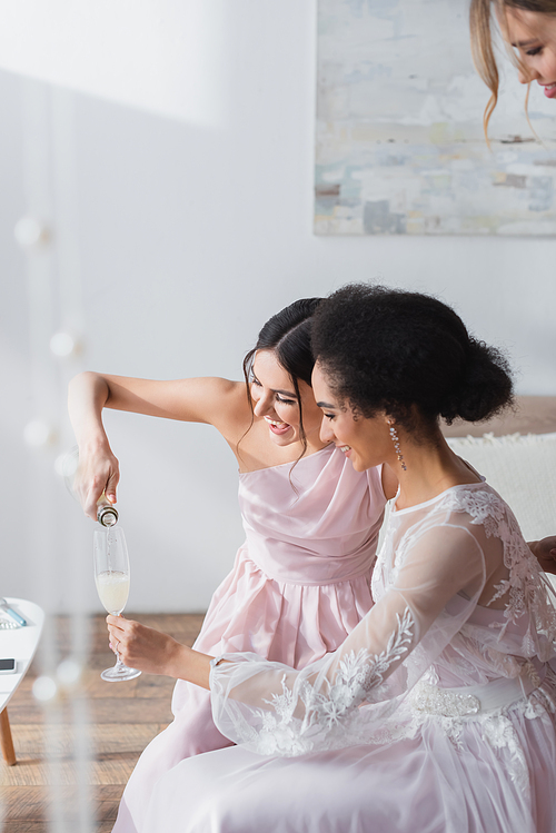 cheerful woman pouring champagne into glass of african american bride in bedroom