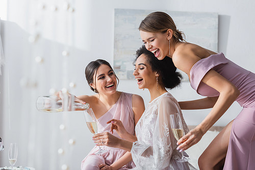 young woman pouring champagne near african american bride and excited bridesmaid