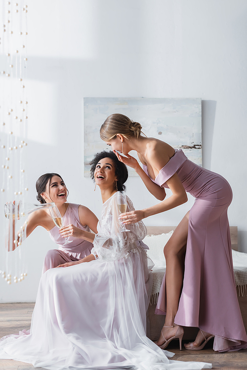cheerful bride with interracial friends talking and drinking champagne in bedroom