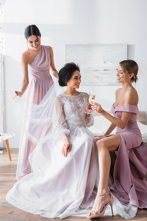 happy african american bride clinking champagne glass with bridesmaid in bedroom