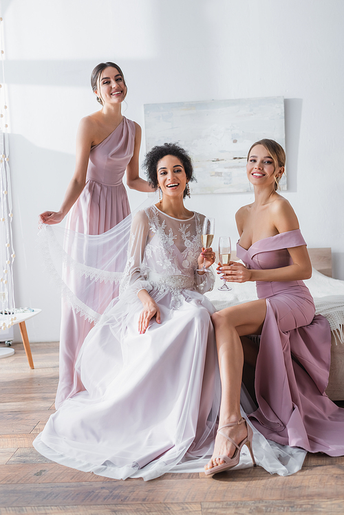 happy african american bride with bridesmaids smiling at camera in bedroom