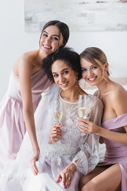 happy bridesmaids smiling at camera while holding champagne near african american bride