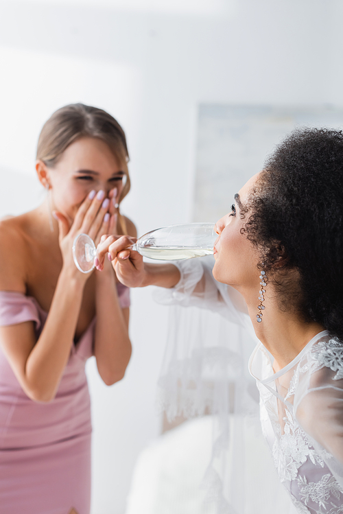 african american bride drinking champagne near laughing friend on blurred background