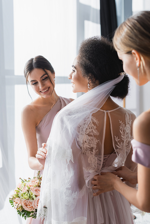 smiling bridesmaids preparing african american bride for wedding, blurred foreground
