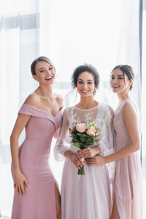 african american woman with wedding bouquet, and cheerful bridesmaids smiling at camera