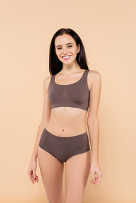 slim woman in underwear smiling at camera while posing isolated on beige