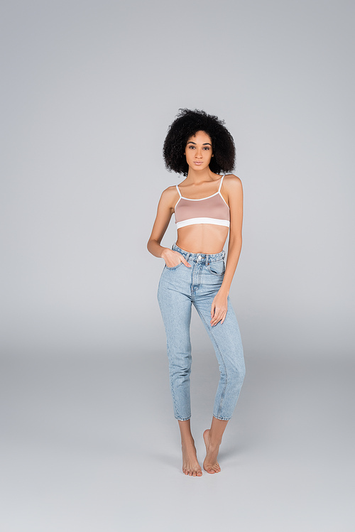 full length view of slim african american woman posing in bra and jeans on grey