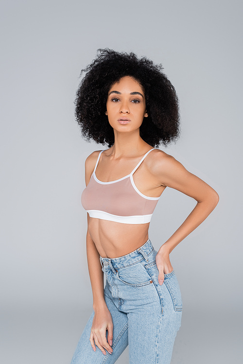 african american woman in jeans and bra posing with hand in back pocket isolated on grey