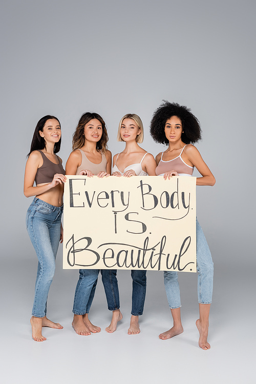 young interracial women holding placard with every body is beautiful lettering on grey