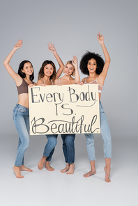 happy multiethnic women holding placard with every body is beautiful lettering on grey