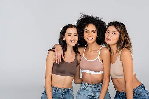 young interracial women in jeans and bras smiling at camera isolated on grey