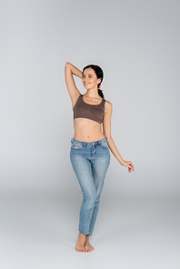 smiling woman in jeans and bra looking away while posing on grey
