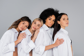 multiethnic women in white shirts leaning on each other while  isolated on grey