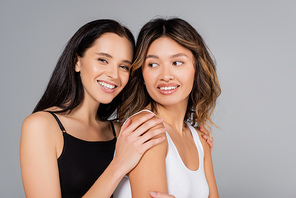 happy woman embracing shoulders of asian friend isolated on grey