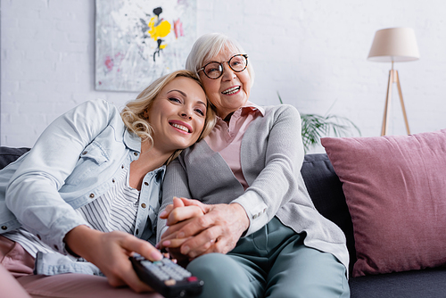 Cheerful senior woman sitting near adult daughter with remote controller on blurred foreground