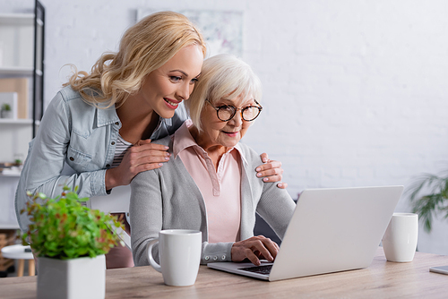 Smiling woman hugging mother using laptop near cups
