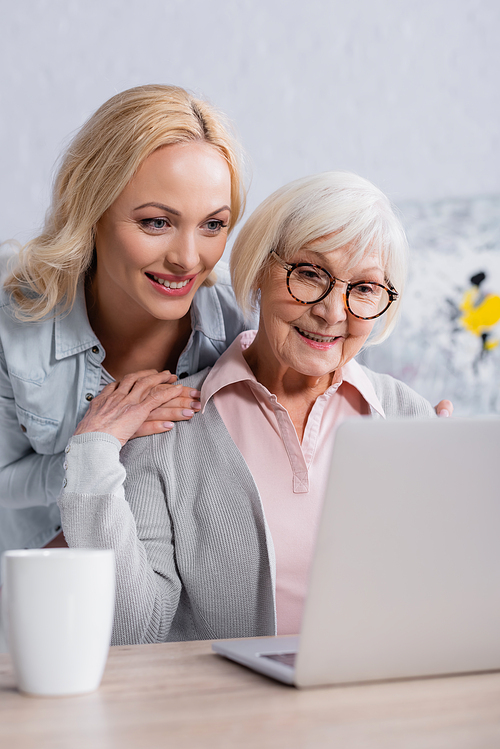 Cheerful woman embracing senior mother near laptop and cup on blurred foreground