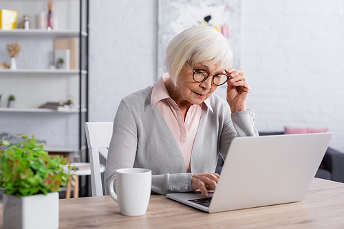 Senior woman in eyeglasses using laptop near cup