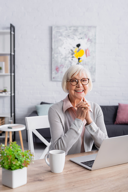 Positive grey haired woman sitting near laptop and cup at home