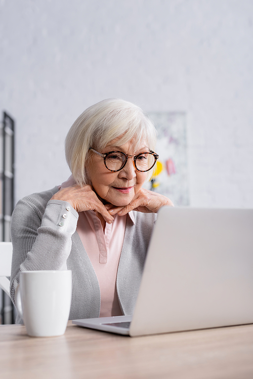 Senior woman in eyeglasses looking at laptop near cup