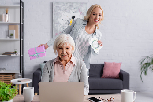 Senior woman using laptop near daughter with present and greeting card on blurred background