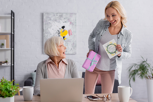 Smiling woman holding greeting card and gift near mother and devices