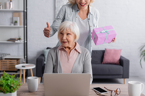 Smiling woman holding present near elderly mother using laptop