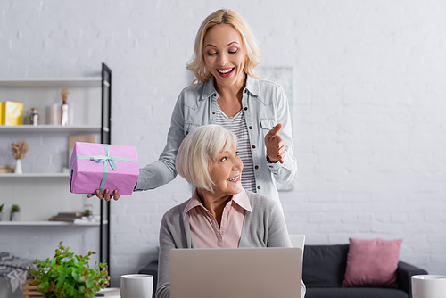 Cheerful woman with gift box standing near senior mother and laptop