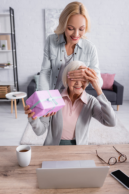 Smiling woman covering eyes to mother while holding present near laptop and tea on table