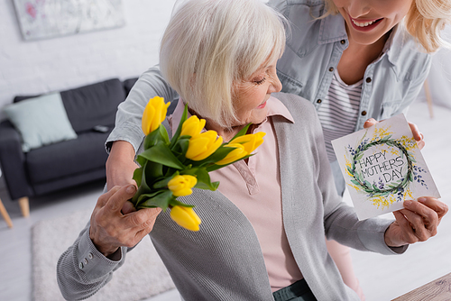 Smiling woman holding greeting card and flowers near senior parent