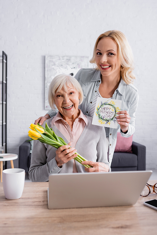 Smiling senior woman holding flowers near daughter with greeting card near laptop
