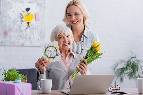 Cheerful woman hugging mother with happy mothers day lettering on greeting card and flowers near laptop