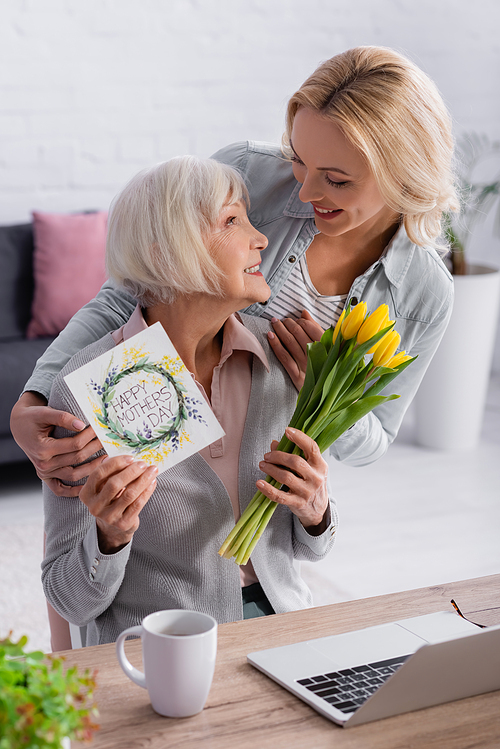Senior woman holding flowers and greeting card with happy mothers day lettering near daughter and laptop