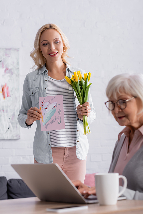 Smiling woman holding greeting card with 8 march lettering and flowers near senior mother using laptop