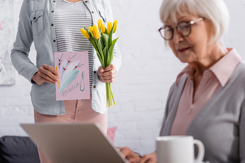 Woman holding tulips and greeting card with 8 march lettering near senior mother using laptop on blurred foreground