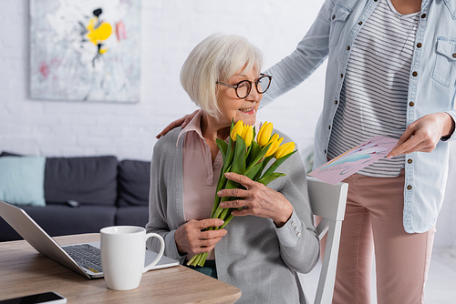 Cheerful elderly woman holding tulips near daughter with greeting card and laptop