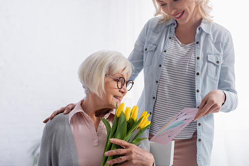 Smiling woman holding greeting card near elderly mother with yellow tulips