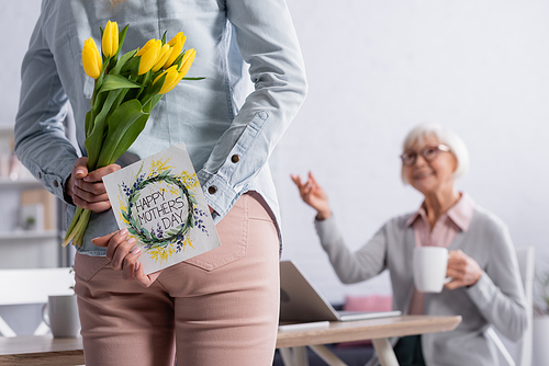 Woman holding greeting card with happy mothers day lettering and flowers near blurred senior mother