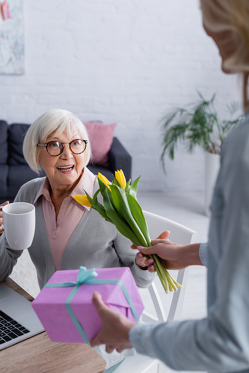 Smiling elderly woman holding cup near daughter with flowers and present