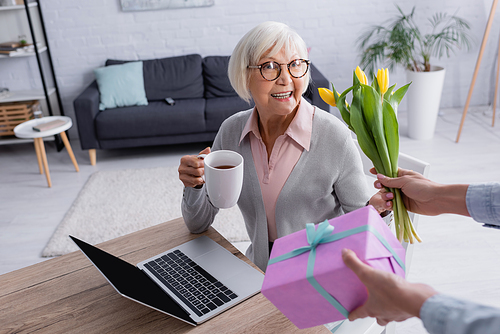 Woman giving flowers and gift box near cheerful mother with tea near laptop
