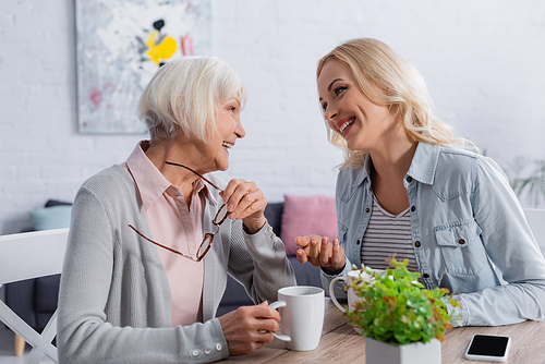 Cheerful woman talking with senior mother near tea and smartphone