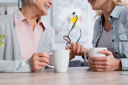 Cropped view of elderly woman holding eyeglasses and cup near smiling daughter