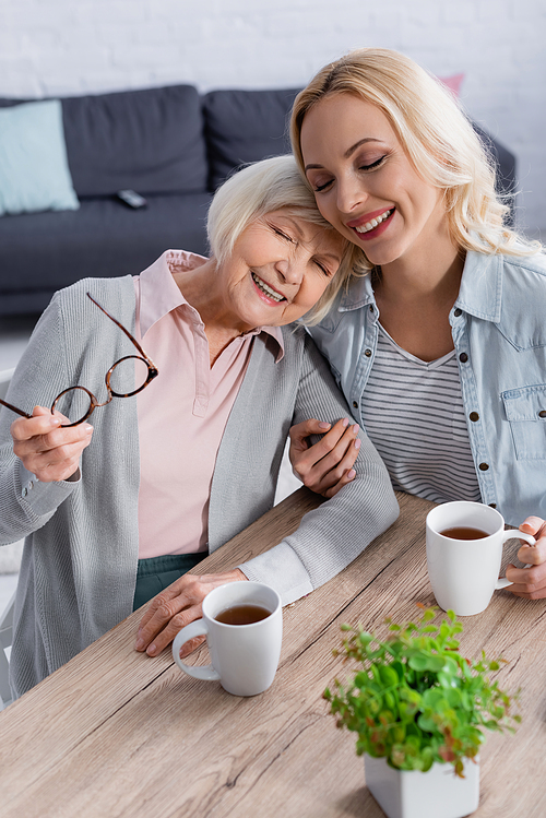 Smiling woman holding cup near elderly woman with eyeglasses