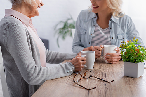 Cropped view of eyeglasses near senior woman holding cup while talking with daughter