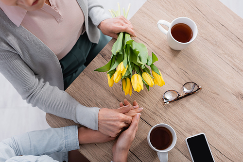 Top view of senior woman with flowers holding hand of daughter near tea and smartphone