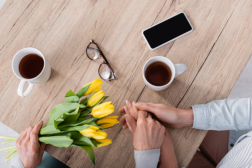Top view of woman holding hand of mother with flowers near tea, eyeglasses and smartphone