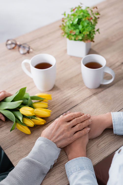 Cropped view of senior woman holding hand of daughter near tulips and cups